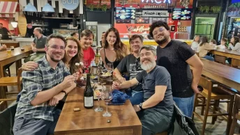Guto e Chai com seu grupo de amigos sorrindo e brindando com vinho em uma mesa do Baxar Mercado em La Plata, capturando a atmosfera animada e acolhedora do local.